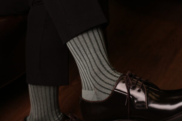 Elegant man wearing light Mint ribbed luxury socks paired with polished dark brown oxford shoes and black trousers, sitting indoors on wooden floor — sophisticated men’s fashion accessory style by Celchuk Luxury Socks.